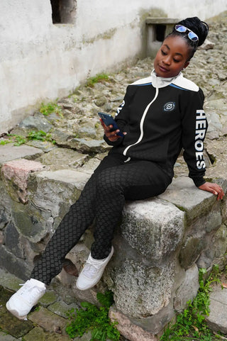 Stylish woman in a black Synergetic Heroes tracksuit sits on a stone ledge, casually looking at her phone. This comfortable and fashionable athleisure set is perfect for everyday wear, workouts, and active lifestyles. Shop Synergetic Heroes for premium apparel today!