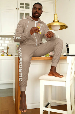 "Stylish man in a beige Synergetic Heroes tracksuit sits on a kitchen counter, casually enjoying a snack. This comfortable and modern athleisure set is perfect for lounging, casual wear, and active lifestyles. Shop Synergetic Heroes for premium apparel!