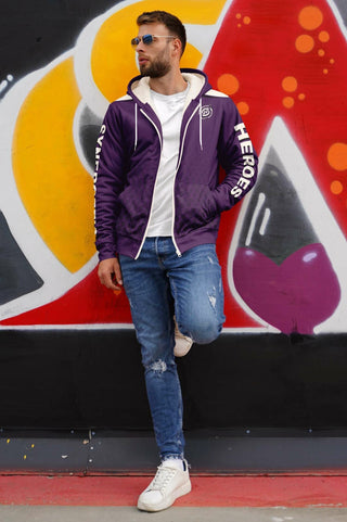 "Stylish man wearing a purple Synergetic Heroes zip-up hoodie, white t-shirt, and ripped jeans, leaning against a colorful graffiti wall. This trendy streetwear look blends comfort and fashion. Shop Synergetic Heroes for premium athleisure apparel today!