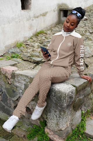 A stylish woman in a beige Synergetic Heroes tracksuit sits on a stone ledge while using her phone. The comfortable athletic wear is perfect for casual and active lifestyles.